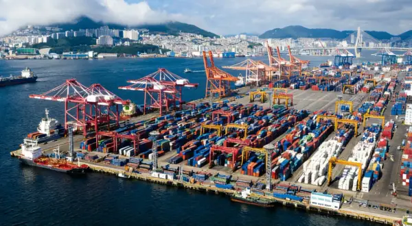 Ogromny port kontenerowy z dźwigami i statkami. W tle widać miasto i most. To skrót do globalnego handlu.