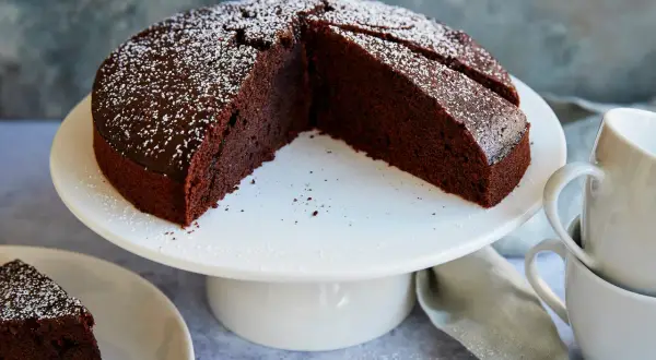 Ein saftiger veganer Schokokuchen, bestäubt mit Puderzucker, steht auf einem weißen Tortenständer. Daneben liegen Tassen und eine Serviette.