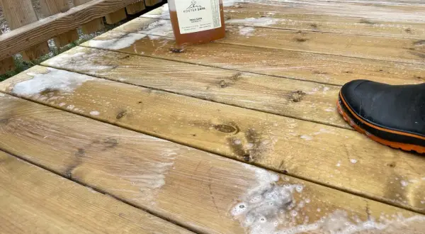 En flaska linoljesåpa står på en altan som håller på att rengöras. En svart stövel med orange kant syns i nedre högra hörnet.