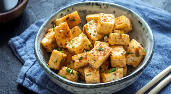 Usmażone kostki tofu, które jest zrobione z ziaren soi, w misce z sezamem i ziołami. Obok pałeczki i sos sojowy.