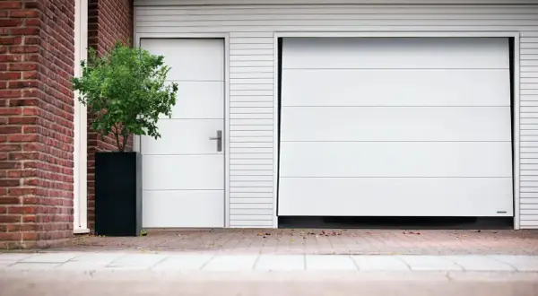 Modernt vipport garage med en vit port och en sidodörr. En grön buske i en svart kruka står bredvid.
