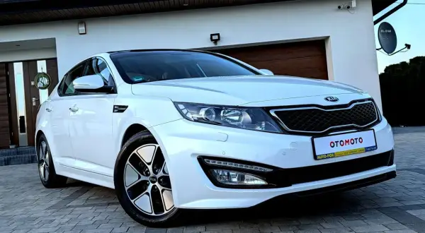 Biały kia sedan z czarnym dachem, zaparkowany przed domem z drewnianą bramą garażową.
