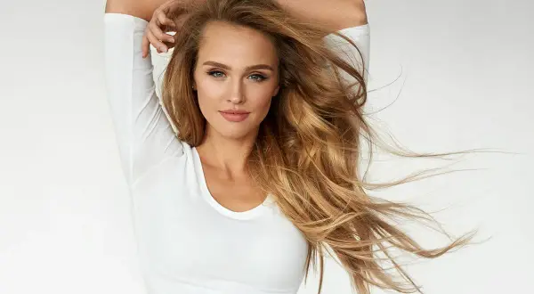 Kobieta z falującymi, beżowymi, ciepłymi blond włosami, w białej bluzce, z rękami uniesionymi nad głową.