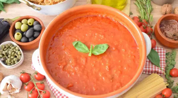 Pyszny, domowy makaron z sosem pomidorowym, przygotowany ze świeżych pomidorów, czosnku i bazylii. Obok składniki do dania: oliwki, kapary, makaron i zioła.