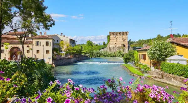 Un incantevole borgo da visitare in Veneto, con un fiume che scorre tra case antiche e una torre medievale. Fiori colorati incorniciano la scena.