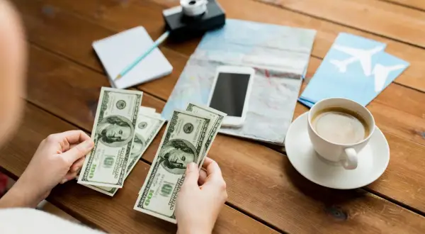 Ręce trzymające banknoty 100 USD, obok mapa, telefon, aparat, notatnik i kawa. Planowanie podróży, a niewypłacanie świadczenia urlopowego co to znaczy?