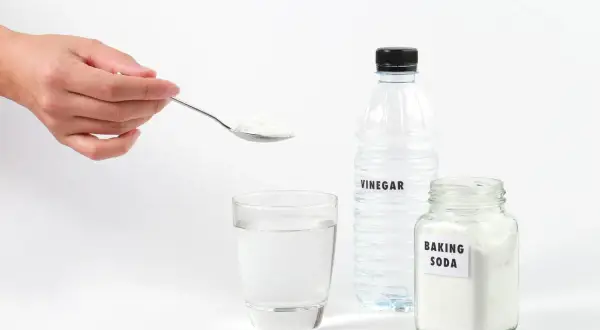 Une main tient une cuillère de bicarbonate de soude pour déboucher une canalisation. Vinaigre et eau sont prêts.
