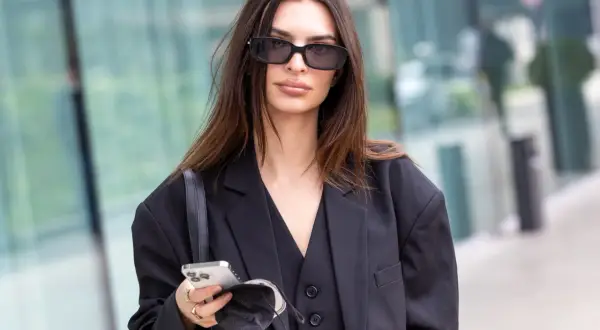 Elegancka brunetka w okularach przeciwsłonecznych, z telefonem i maseczką w dłoni. Jej włosy mają piękny, ciemny kolor.