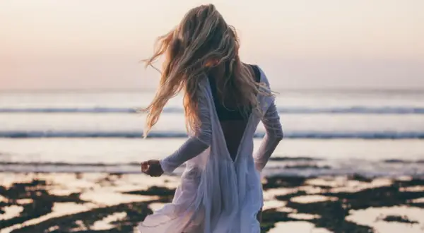 Blondynka w zwiewnej białej sukience tańczy na plaży, a wiatr rozwiewa jej włosy. W tle widać spokojne beach waves i zachodzące słońce.