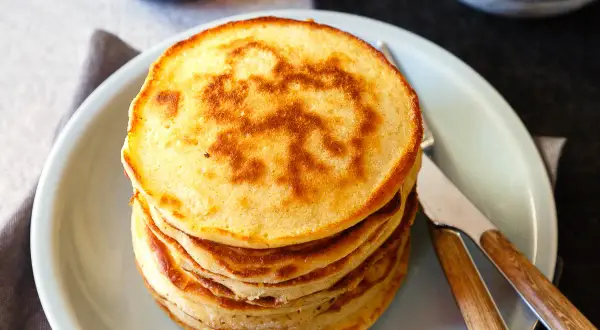 Stapel goldbrauner Dinkel Pancakes mit einem Muster auf dem obersten Pancake, serviert auf einem hellblauen Teller mit Messer und Gabel.