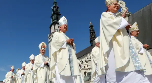 Biskup w białej szacie i mitrze z Matką Boską, na tle wieży kościelnej.