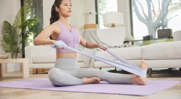 Kobieta ćwiczy pilates w domu, siedząc na macie i napinając taśmę oporową.