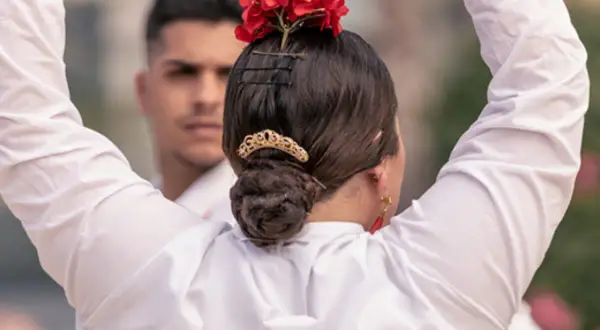 Peinados para bailar flamenco: moño bajo adornado con flores rojas y peineta dorada. Un hombre con camisa blanca al fondo.