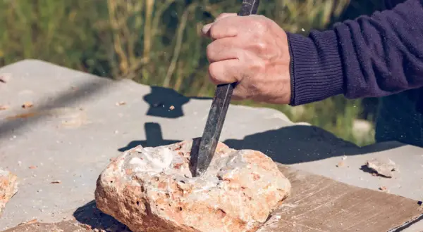 Dłoń z dłutem pracuje nad kamieniem, tworząc otwór. To precyzyjne wiercenie w granicie, gdzie każdy uderzenie kształtuje materiał.
