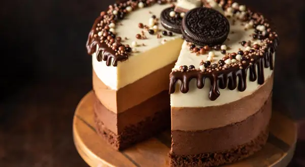 Torta ai tre cioccolati ricetta facile: golosa e scenografica, con strati di mousse al cioccolato bianco, al latte e fondente, decorata con gocce di cioccolato e biscotti.