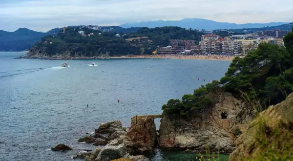Widok na zatokę z łodziami i plażą. W oddali widać miasto i góry. Pogoda na Costa Brava zapowiada się na spokojną.