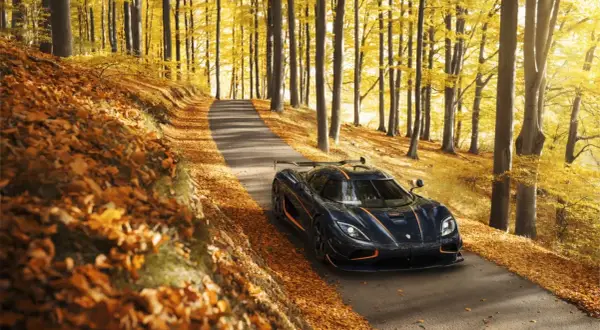 Mroczny Koenigsegg Agera RS z pomarańczowymi akcentami przemierza jesienną drogę wśród złotych liści i drzew.