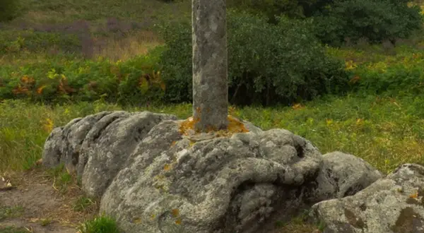 Pedra da serpe, un cruceiro de pedra incrustado nunha rocha con forma de serpe, rodeado de vexetación verde e púrpura.