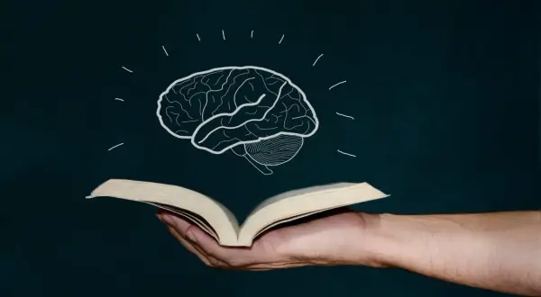 Mano sostiene libro abierto, de él surge un cerebro dibujado, símbolo de la lectura global y el conocimiento.