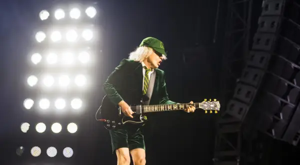 Angus Young von AC/DC spielt auf der Bühne, während die Scheinwerfer leuchten. Seine ikonische Kleidung und die Gitarre lassen an bekannte Lieder denken.