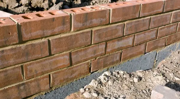 Ceglana ściana ogrodzenia z widocznym szarym bloczkiem fundamentowym, który stanowi alternatywę dla tradycyjnej podmurówki.
