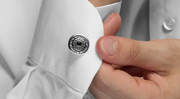 Dettaglio su come si mettono i gemelli: una mano inserisce un gemello rotondo con decorazioni argentate e una pietra nera in un polsino di camicia bianca.