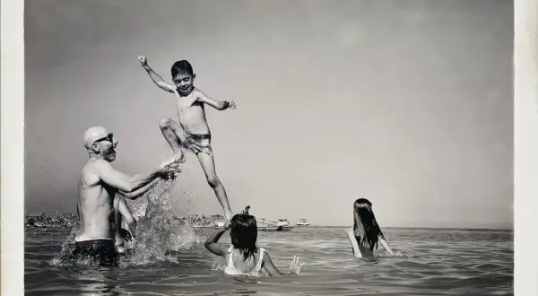 Un bambino salta in aria, sorretto da un uomo con occhiali da sole, mentre due bambine nuotano. Un momento di gioia estiva, un ricordo di Carlo Traini.