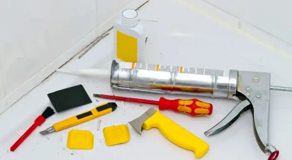 Werkzeuge zum Silikon von Fliesen entfernen: Kartuschenpresse, Cutter, Schaber, Bürste und eine Flasche mit Reiniger.