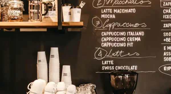 Tafel mit Kaffeebar Ideen: Macchiatos, Cappuccinos, Lattes und mehr. Stapel von Pappbechern und Espressomaschine im Vordergrund.
