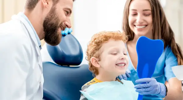 Uśmiechnięty chłopiec ogląda swoje zęby w lusterku w kształcie zęba, podczas gdy dentysta i asystentka patrzą z aprobatą. Czy fluoryzacja jest szkodliwa? Nie, to zabieg chroniący zęby.