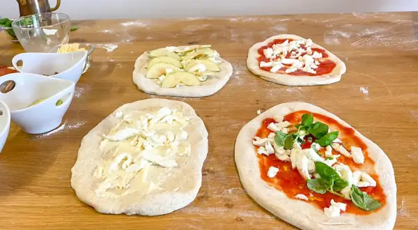 Vier rohe Pinsa-Teige auf einem Holztisch, teils belegt mit Tomatensoße, Mozzarella, Äpfeln und Basilikum.