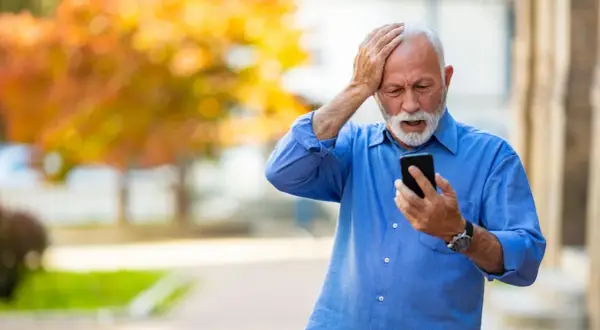 Anziano con mano sulla testa, guarda il telefono con espressione preoccupata, forse per la paura di parlare al telefono.