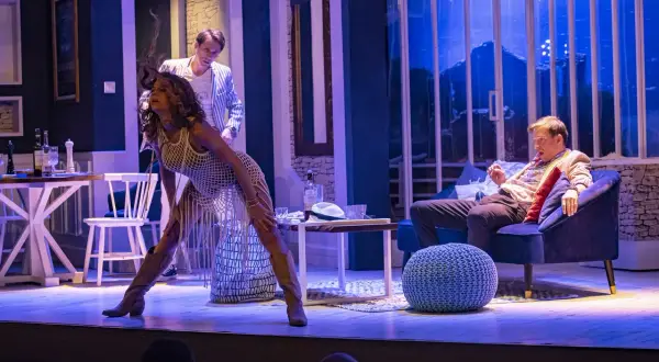 Scena teatralna: kobieta w białej, ażurowej sukience i mężczyzna w marynarce siedzą na kanapie.