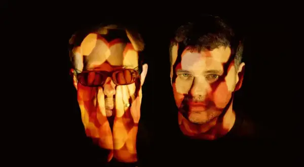 The Chemical Brothers logo is projected onto the faces of two men, one wearing sunglasses, in a dark, atmospheric setting.