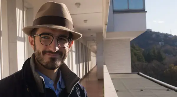 Ruggero Rollini, con cappello e occhiali, sorride su un balcone con vista su un paesaggio collinare.