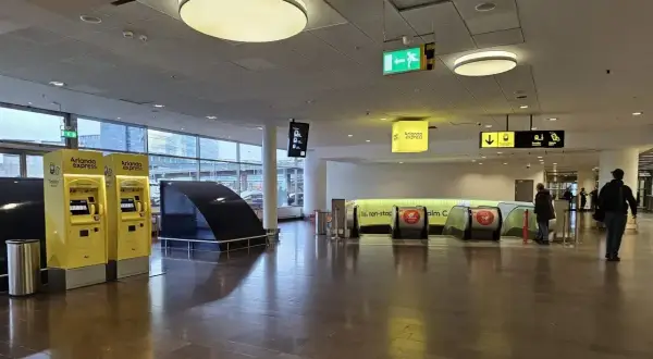 Dwie żółte kasy biletowe Arlanda Express, gdzie sprawdzisz arlanda express cena. Dalej schody ruchome i ludzie.