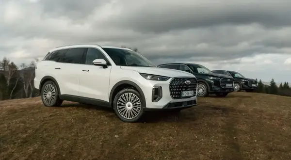 Biały Jaecoo 9 i dwa ciemne SUV-y na wzgórzu pod pochmurnym niebem.