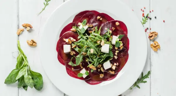 Rote Bete Carpaccio mit Rucola, Walnüssen und Feta auf einem weißen Teller, garniert mit frischem Basilikum.