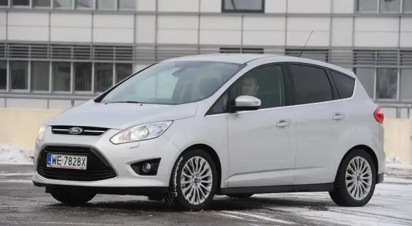 Srebrny Ford C-Max z włączonymi przednimi światłami, gotowy do jazdy w zimowych warunkach.