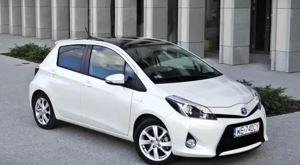 Biały Toyota Yaris z czarnym dachem, zaparkowany przed nowoczesnym budynkiem. Wiele osób szuka takich aut, czytając toyota yaris opinie.