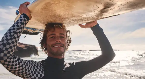 Lächelnder Surfer mit lockiger Surfer-Frisur trägt Neoprenanzug und hält Surfbrett über dem Kopf.