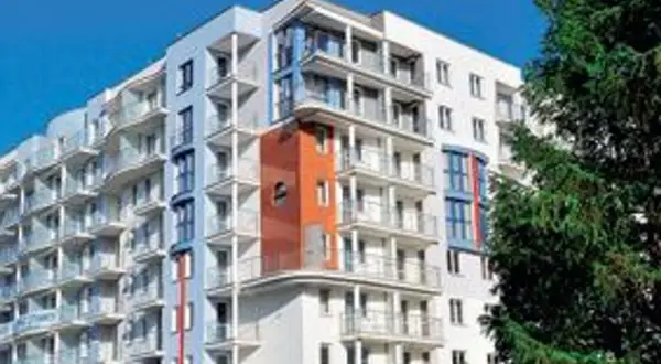 Nowoczesny hotel z balkonami i pomarańczowym akcentem architektonicznym, na tle błękitnego nieba i drzew.