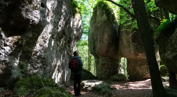 Ein Wanderer steht in einer **steinernen Stadt** aus Felsformationen, umgeben von üppigem Grün und Sonnenlicht.
