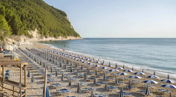 Filettata di ombrelloni e lettini sulla spiaggia di San Michele, con il mare azzurro e la collina verde sullo sfondo.