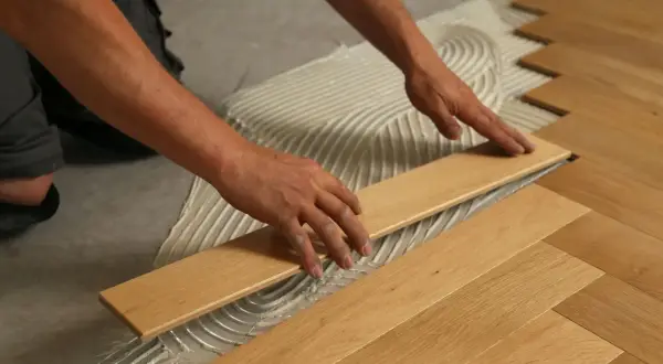 Manos colocando tablones de maderas duras sobre adhesivo para crear un suelo de espiga.