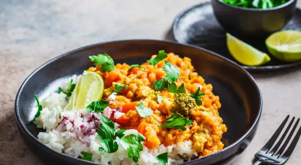 Un plat de riz blanc garni d'un curry de lentilles corail et légumes, parsemé de coriandre fraîche et de quartiers de citron vert. Une recette d'été parfaite.