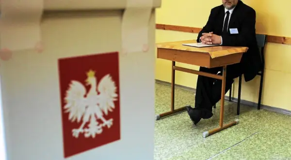 Mąż zaufania w garniturze siedzi przy biurku w lokalu wyborczym, obok urny z godłem Polski.
