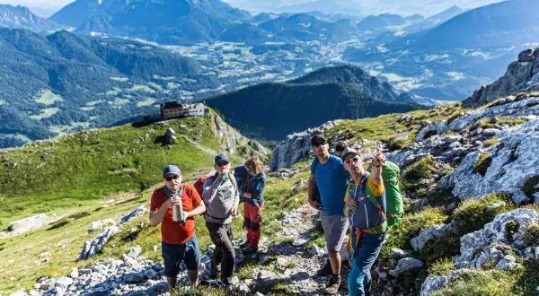 Wandergruppe auf dem Weg zum hocheck Watzmann. Im Hintergrund ein Alpenpanorama mit einem Dorf.