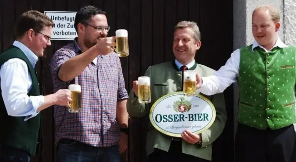 Vier Männer stoßen mit Biergläsern an. Einer hält ein Schild der Osser Brauerei.