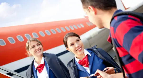 Zwei lächelnde Flugbegleiterinnen im blauen Anzug mit roten Halstüchern prüfen eine Bordkarte vor einem Flugzeug. Die Ausbildung zur Flugbegleiterin und das Gehalt sind attraktiv.